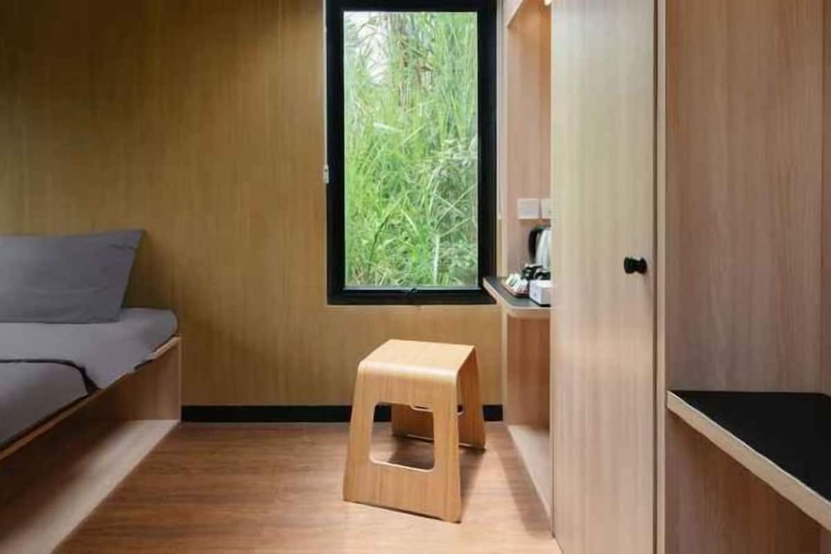 Una habitación con cama, un taburete, una ventana con vistas a la vegetación y paredes de madera.