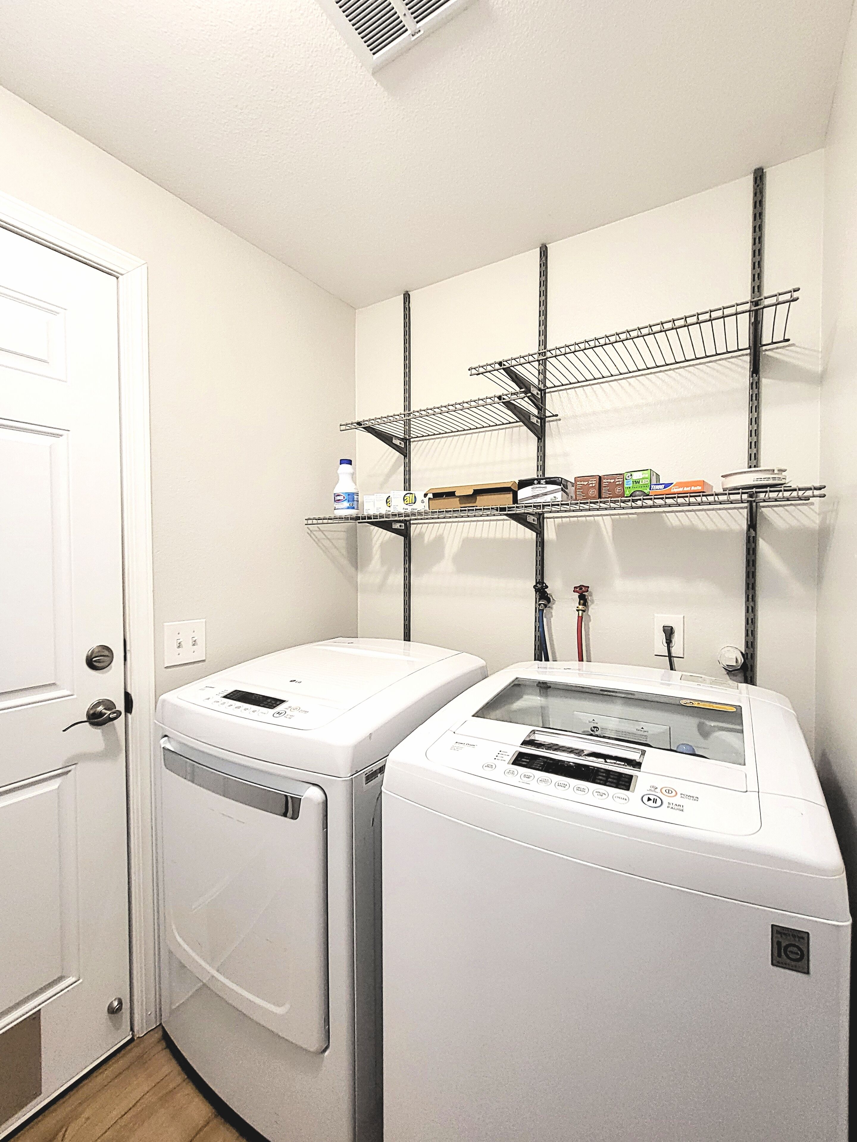 A laundry room with two front-loading washing machines, a metal shelf with various items, and a white door.