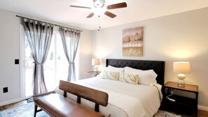 Ein Schlafzimmer mit Bett, Sitzbank, Deckenventilator, Nachttischen, Lampen und einem großen Fenster mit Vorhängen.