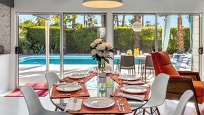 Espaço de refeições com vista para uma piscina, mesa posta para refeição e uma grande luminária pendente.