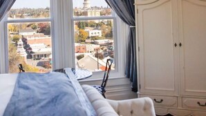 A bedroom with a large window, a bed, a wardrobe, and a view of a cityscape.