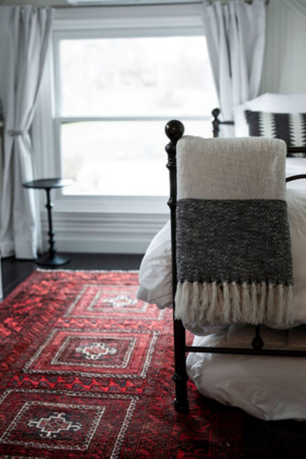 A bed with a white and gray blanket, a red patterned rug, and a window with curtains.