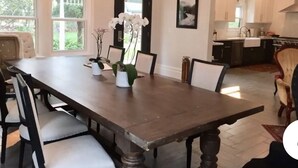 A dining room with a large wooden table, chairs, and a chandelier. There is a kitchen visible in the background.