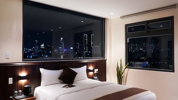A hotel room with a large bed, two bedside tables, a nightstand, a telephone, and a view of a cityscape at night. - Bros Hotel (Ho Chi Minh City)