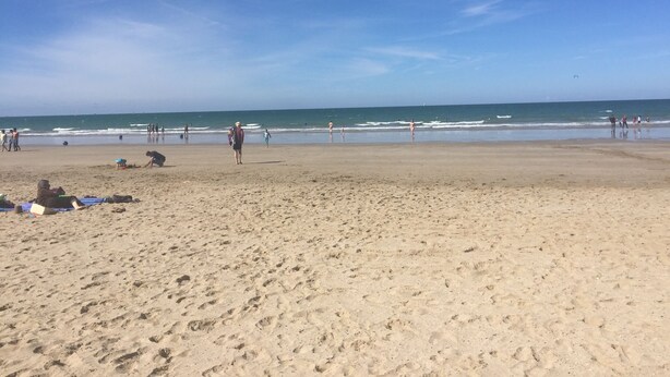 Une vaste plage de sable fin parsemée d’empreintes, un ciel bleu azur et une mer calme.