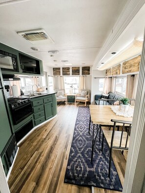 A compact kitchen with a microwave, stove, and sink. There is a dining area with a table and chairs. - RustiCamper Near St. George and Zion (Leeds)