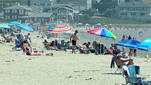 Una playa con gente tomando el sol y nadando, sombrillas y casas al fondo.