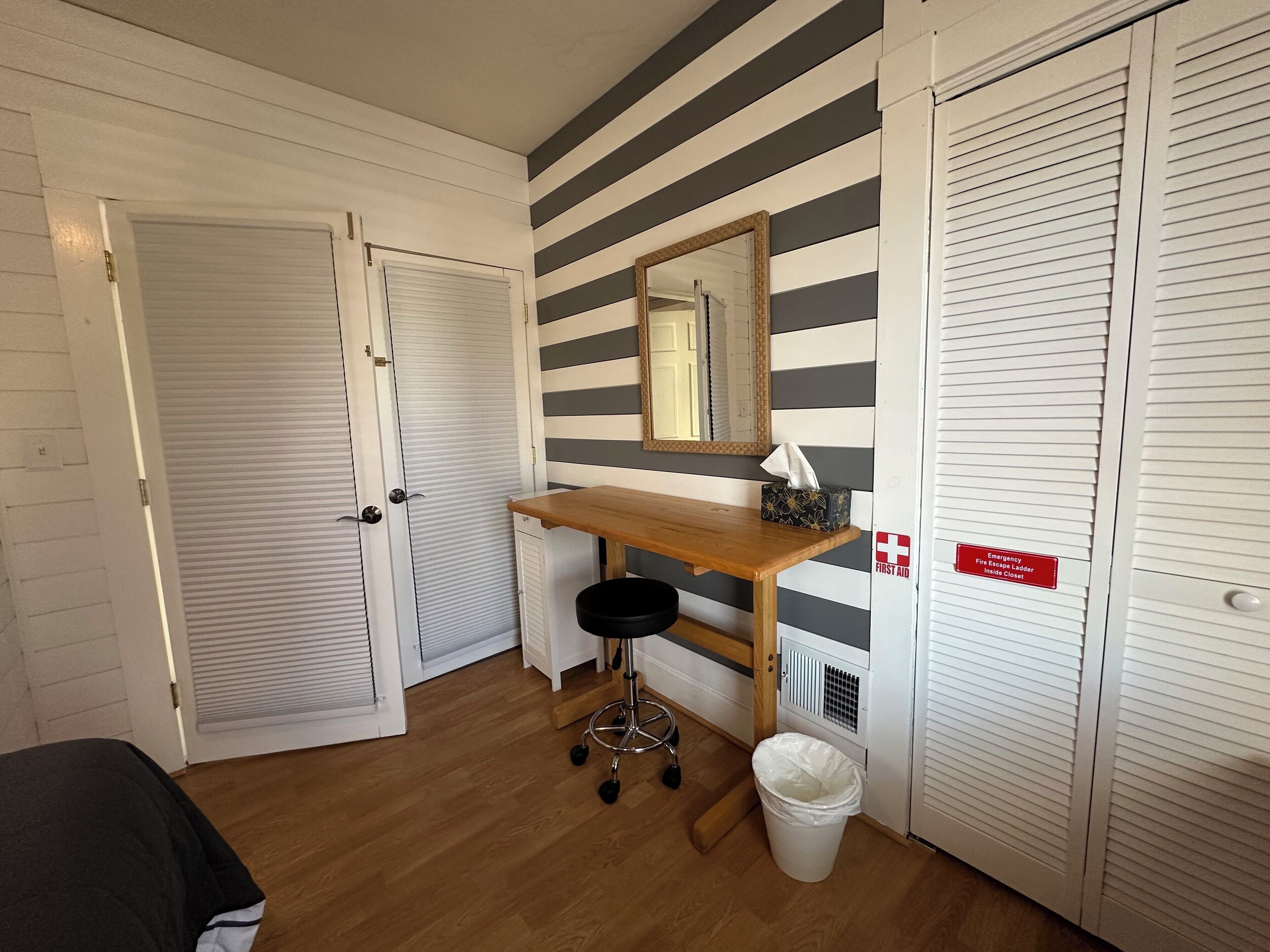 A small room with a wooden desk, a black office chair, a white trash bin, and a mirror on the wall. There is a door on the left and a closet on the right.