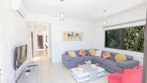 A modern living room with a grey sofa, a white coffee table, and a pink chair. There is a flat-screen TV on a white stand, and a painting on the wall. - Protaras Holiday Villa My5 (Protaras)