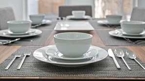 Uma mesa de jantar posta para quatro pessoas, com tigelas e pratos brancos, talheres e um jogo americano trançado.