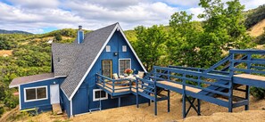 A blue house with a balcony, a wooden deck, and a view of a hilly landscape.