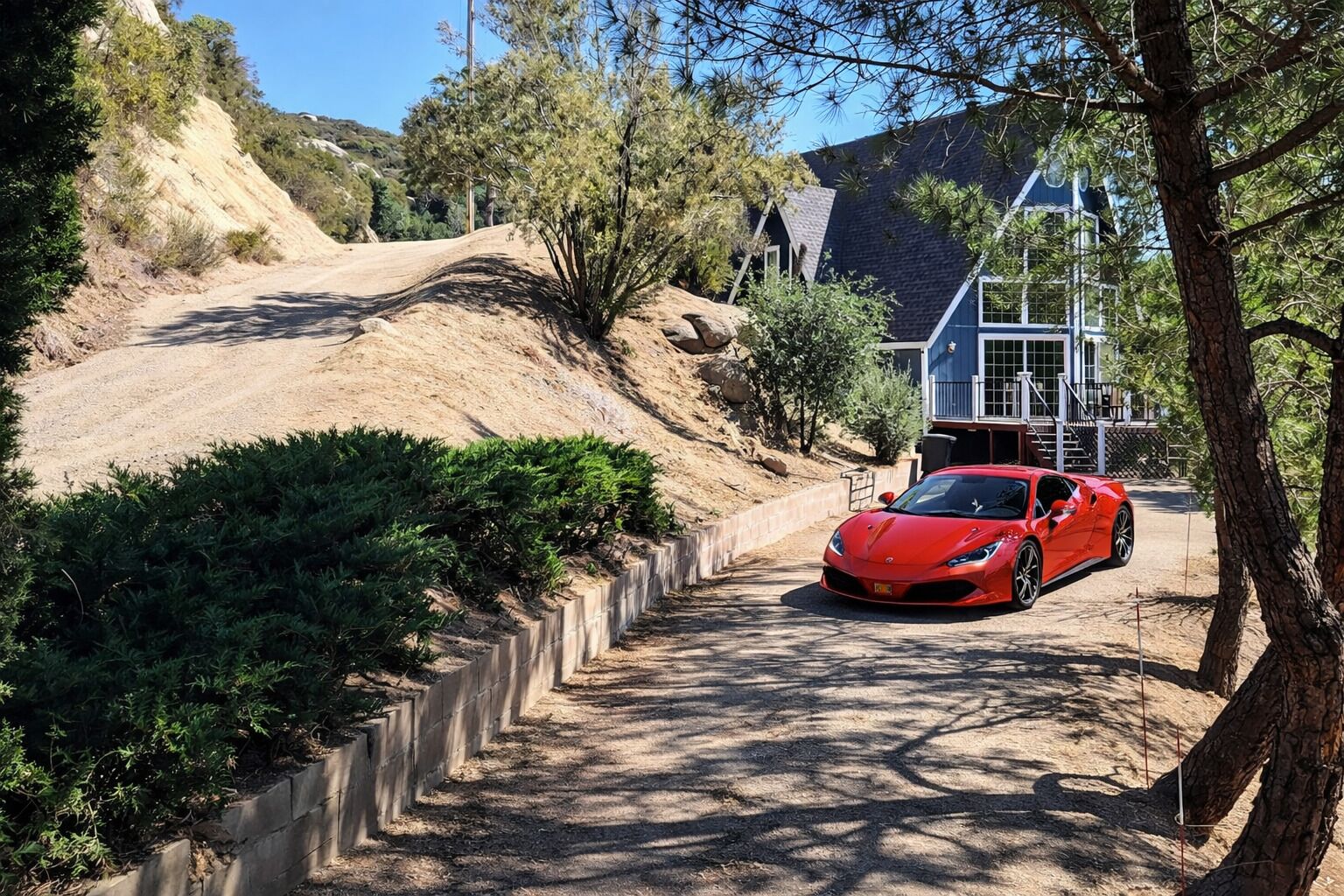 A red sports car parked on a dirt path beside a house with a stone retaining wall and greenery.