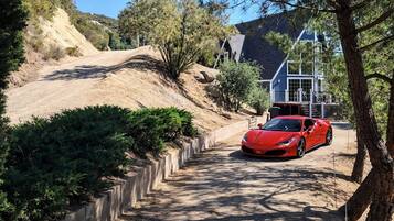 A red sports car parked on a dirt path beside a house with a stone retaining wall and greenery.