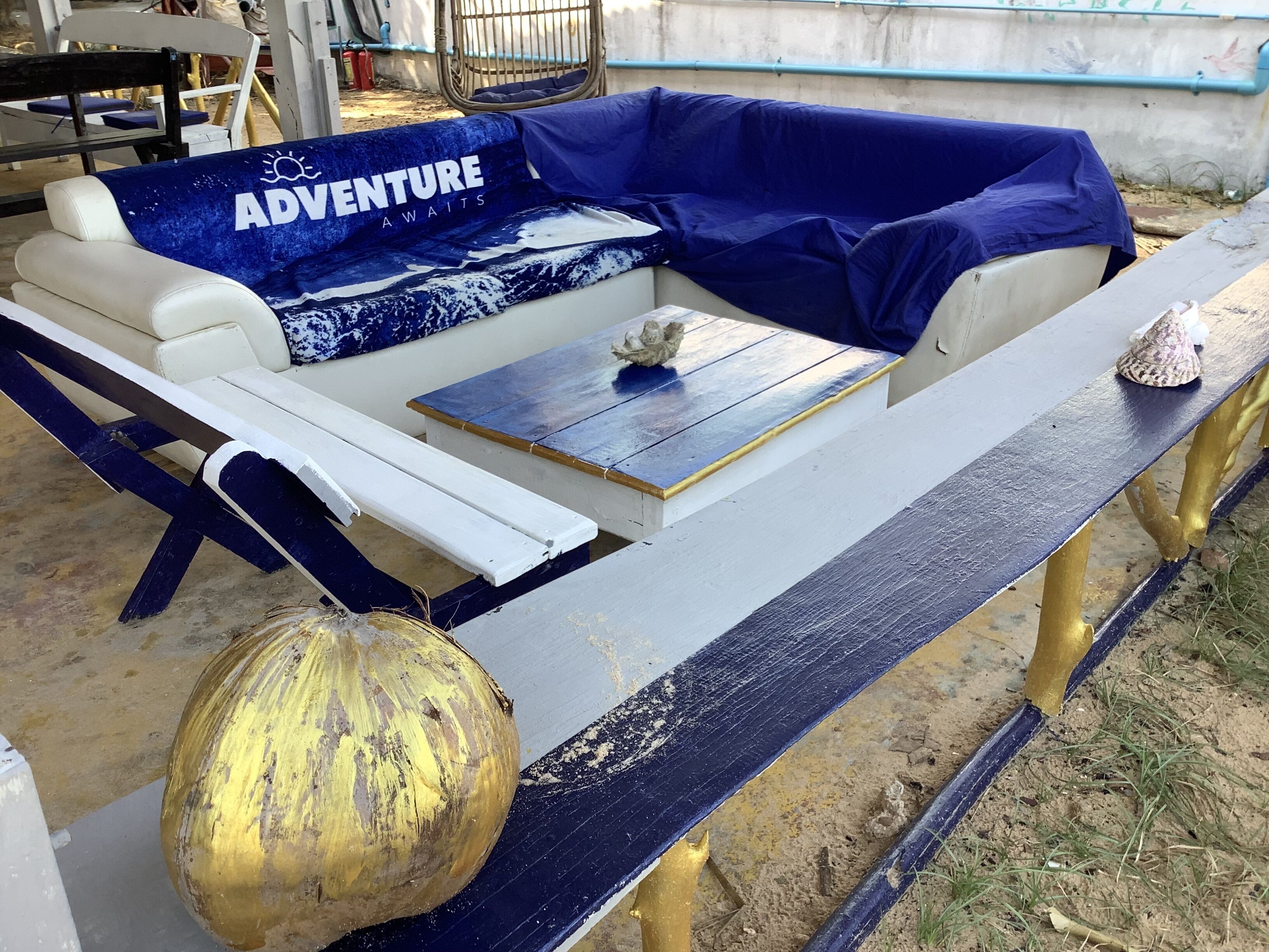 A blue and white outdoor seating area with a 'Adventure Awaits' sign, a table, and a decorative shell.