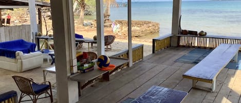 Un espacio de estar techado al aire libre con vista al mar, que incluye un sofá, sillas y una mesa con una pelota de voleibol.