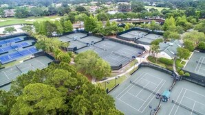 Vista aérea de un complejo de canchas de tenis con varias pistas, rodeado de árboles y edificios.