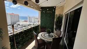Un balcón con una mesa redonda y sillas, luces colgantes, una pared verde y vistas a la ciudad y al océano.