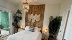 Un dormitorio con una cama grande, cabecera de madera, decoración de pared con macramé, lámparas de noche y vistas al exterior con vegetación.