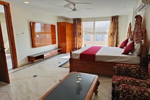 A hotel room with a bed, wooden wardrobe, a ceiling fan, a large window with curtains, a small table with a glass, and a patterned armchair. - Hotel Right Palace And Banquet (New Delhi)