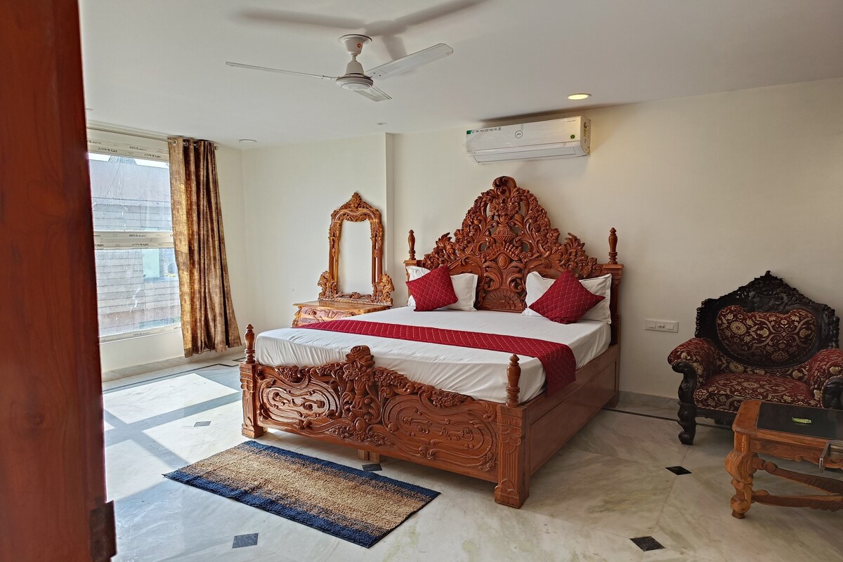 A spacious bedroom with a large bed, a ceiling fan, an air conditioning unit, a wooden chair, and a view of the outside through a window with curtains.