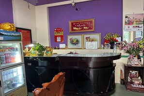 A reception area with a purple wall, a counter with framed certificates, a refrigerator, and decorative plants. - Thuy Duong Hotel Binh Duong (Ho Chi Minh City)