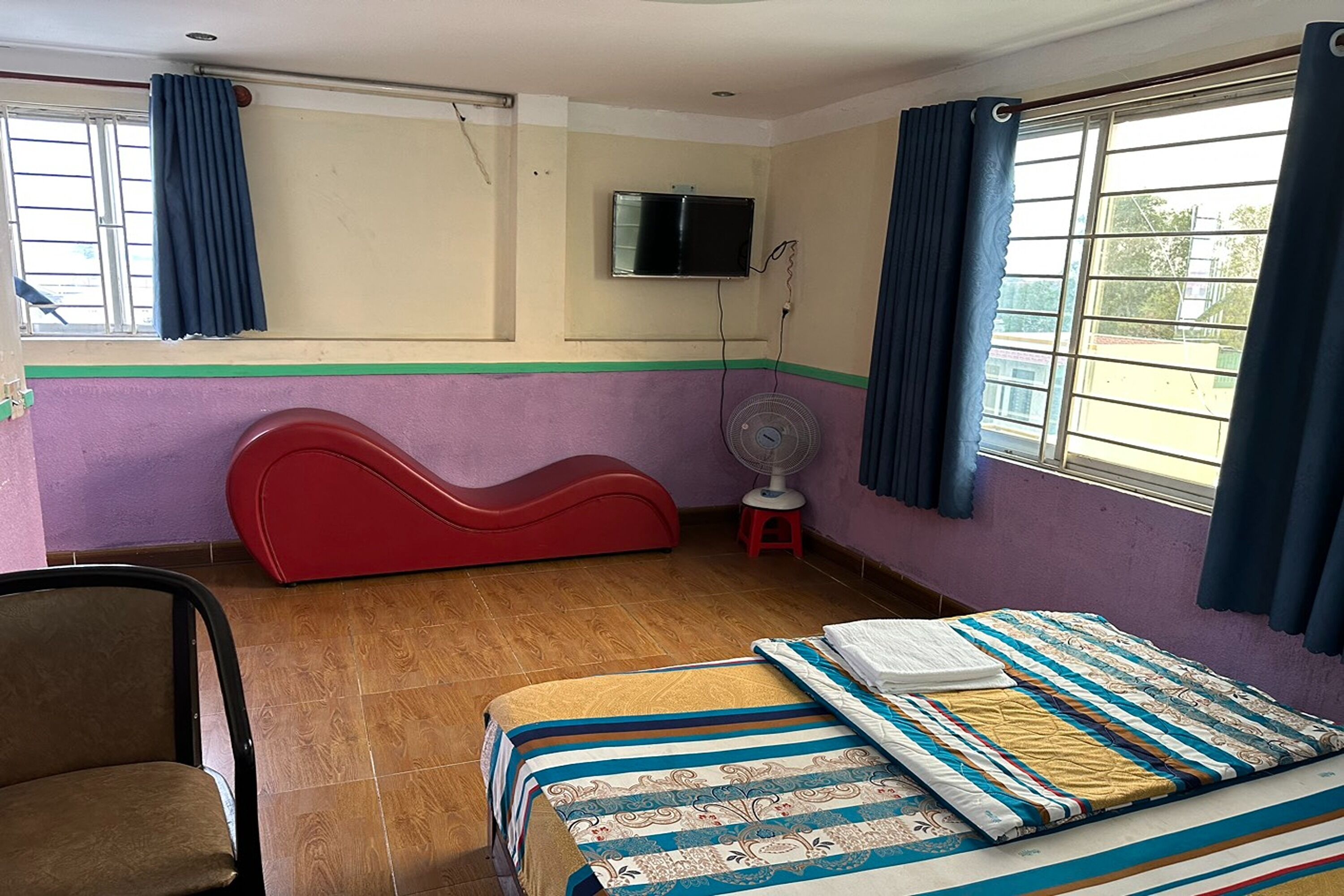 A room with a bed, a red couch, a chair, a TV mounted on the wall, and a window with curtains.