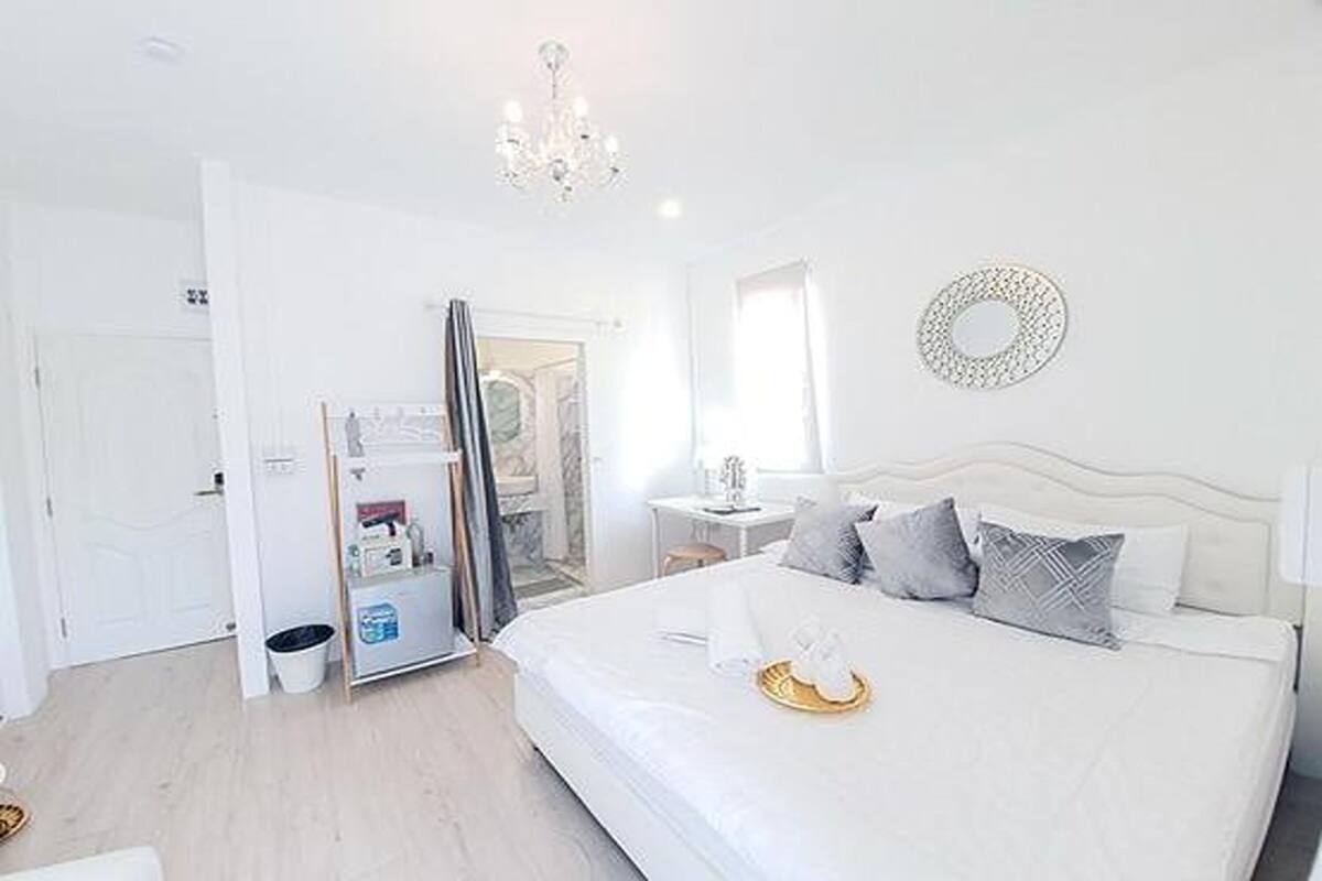 A bright, white hotel room with a large bed, a chandelier, and a view of the outside.