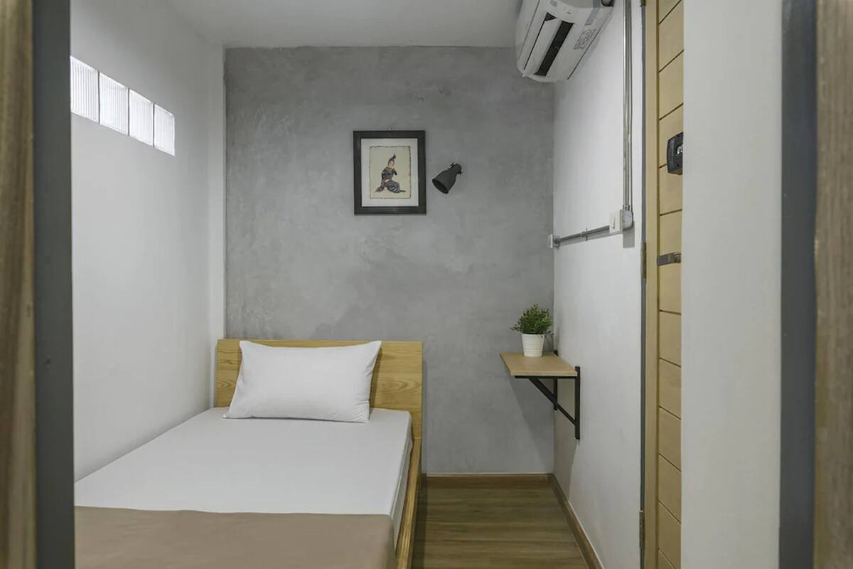 Een kleine, smalle kamer met een bed, een klein plankje met een plant, een ingelijst beeld aan de muur en een raam met zonwering.