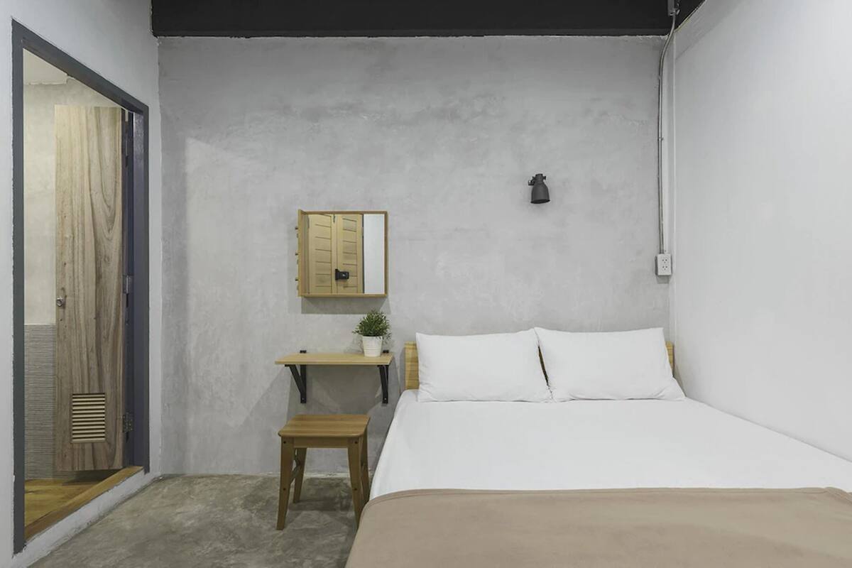 Een slaapkamer met een bed, bureau, stoel, een klein tafeltje, een plant in pot en een spiegel aan de muur.