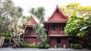 Un bâtiment traditionnel à toit rouge, doté d’un balcon, entouré d’une végétation luxuriante et orné d’une petite statue de chien à l’entrée.