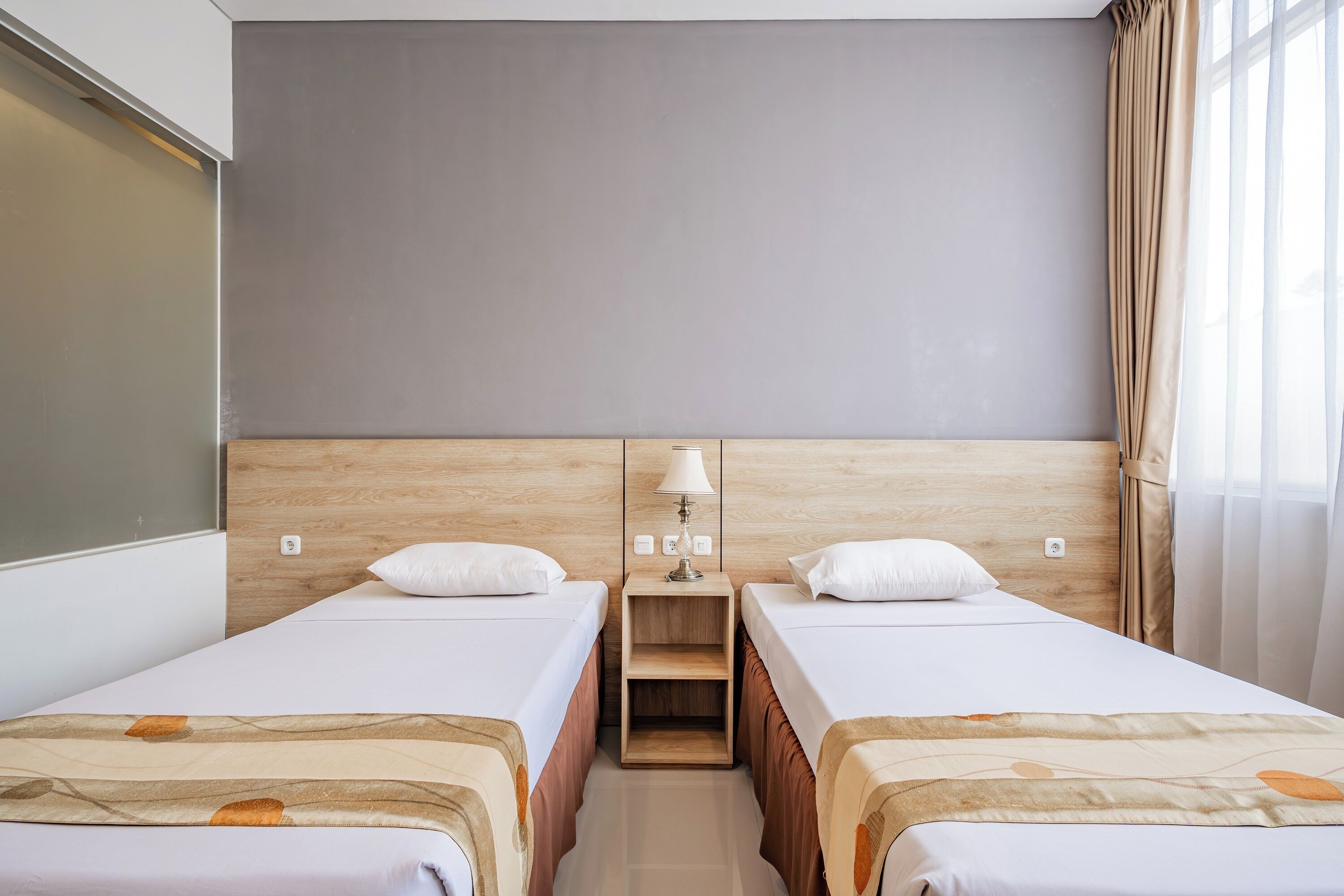 A hotel room with two single beds, a nightstand with a lamp, and a window with curtains.