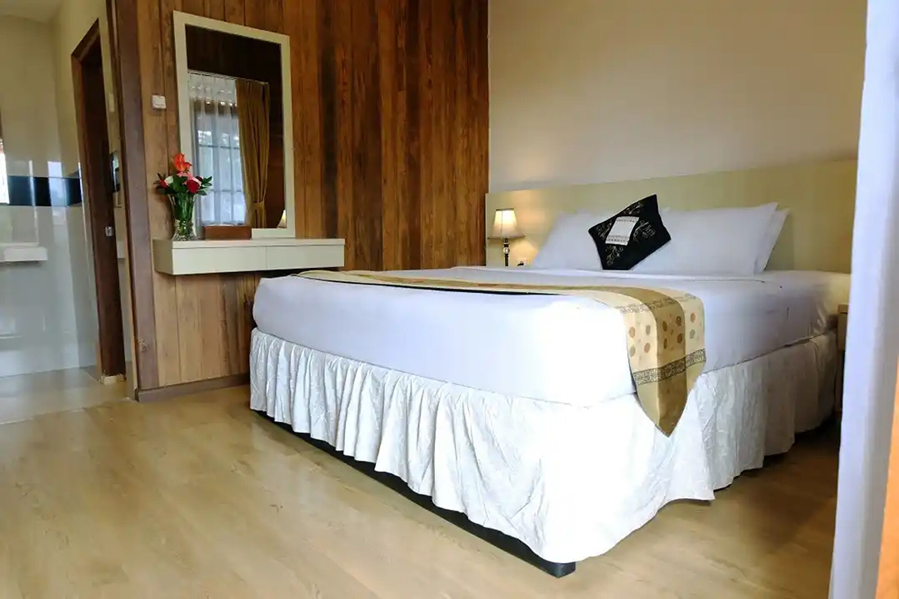 Une chambre d’hôtel avec un grand lit, des boiseries, un miroir, une table de chevet avec une lampe et un vase rempli de fleurs.
