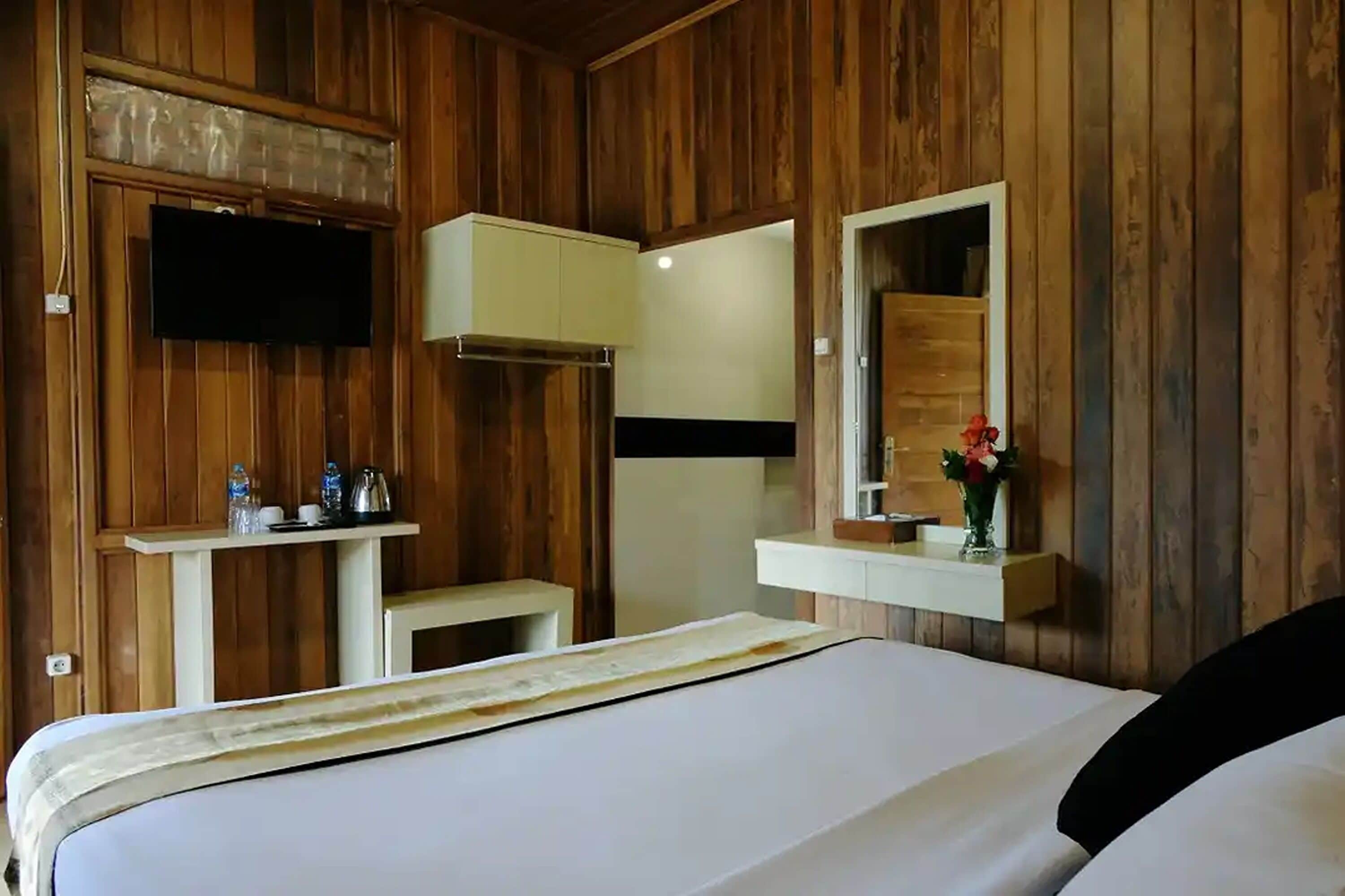 A wooden-paneled room with a bed, a TV mounted on the wall, a small table with a mirror, and a shelf with toiletries.