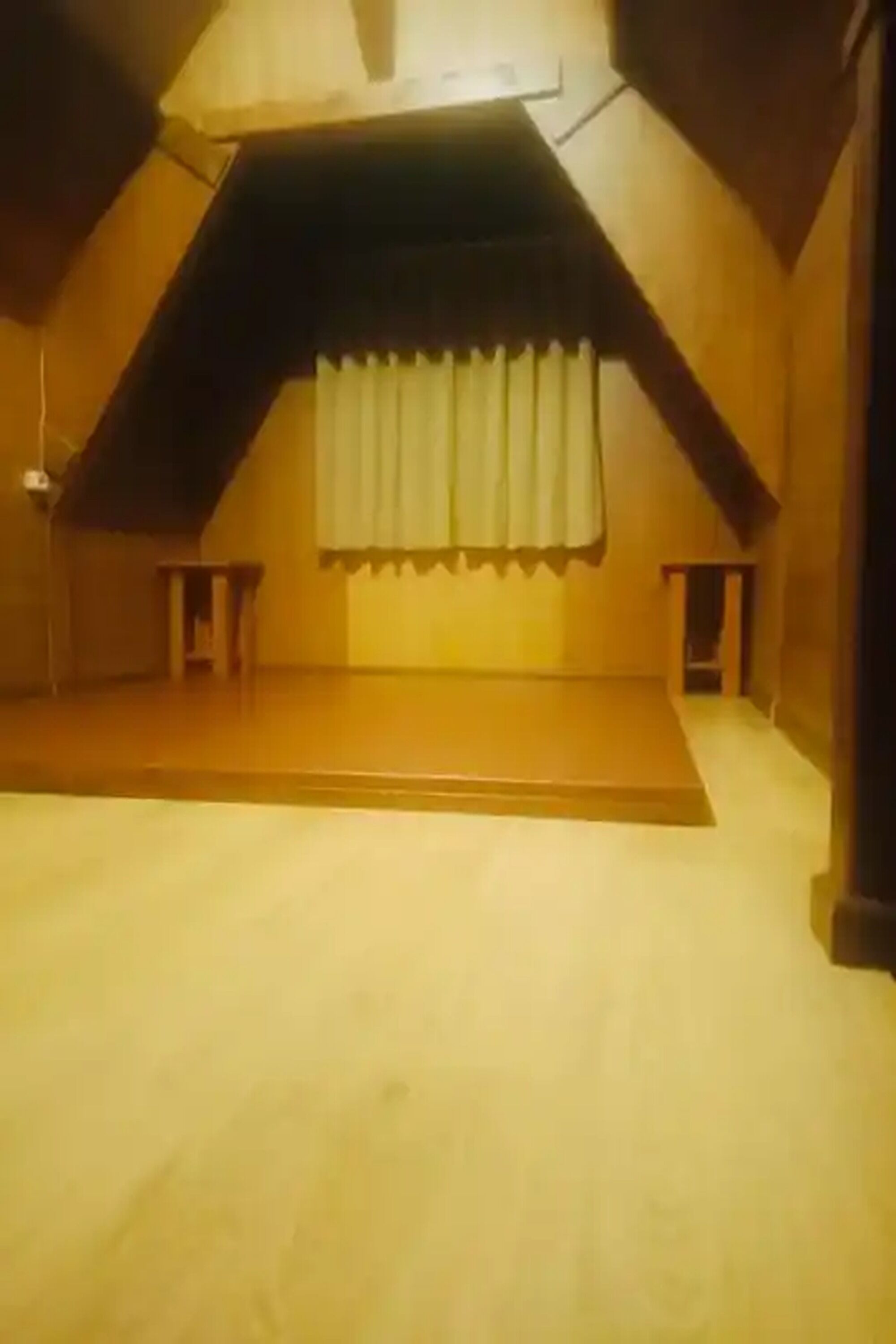 Une pièce avec un plafond en pente, une petite fenêtre avec un rideau et un plancher en bois.
