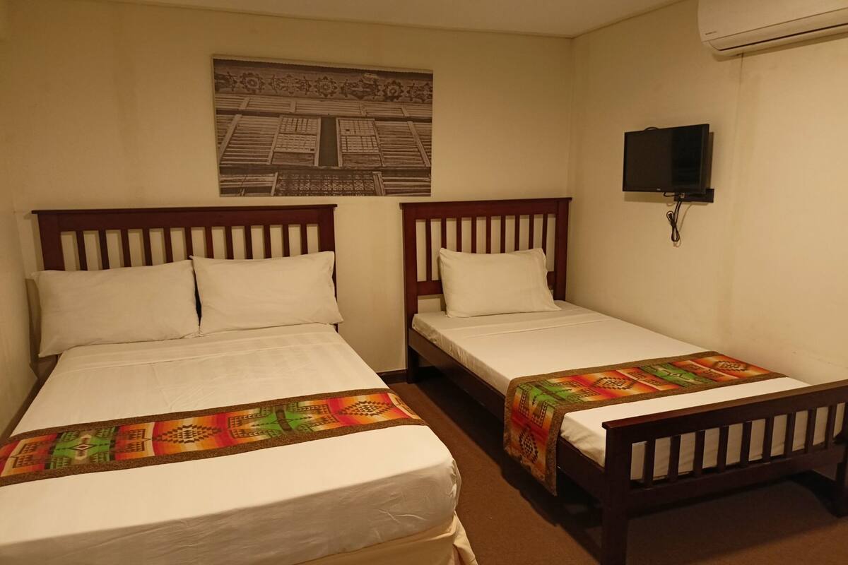 A hotel room with two single beds, a television mounted on the wall, and a decorative wall hanging.