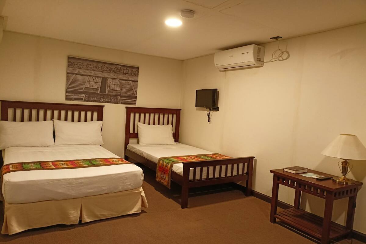 A hotel room with two single beds, a small table with a lamp, an air conditioning unit, and a television on the wall.