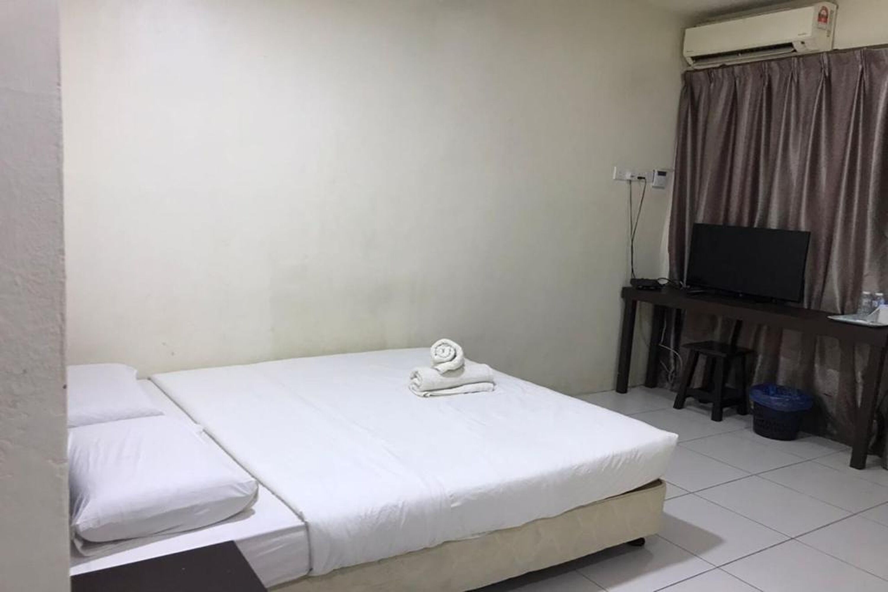 Una cama individual con ropa blanca y una toalla enrollada, un televisor sobre un soporte de madera y un equipo de aire acondicionado en la pared.