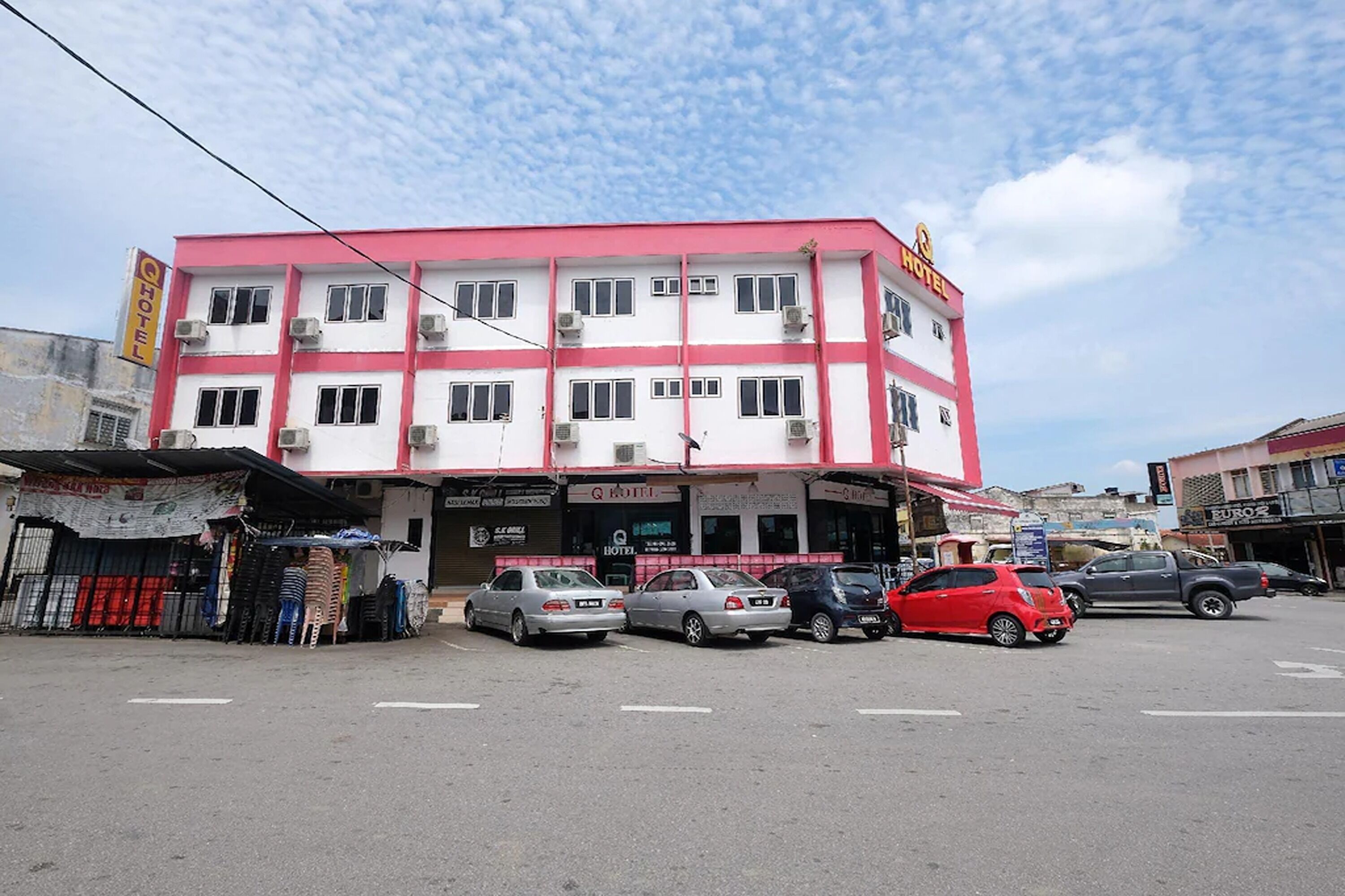A multi-story hotel with a pink and white facade, several parked cars, and a clear sky.
