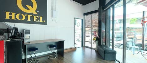 A hotel reception area with a large sign reading "Q HOTEL," a reception desk, two bar stools, a small kitchenette, and a large glass door leading to an outdoor seating area.
