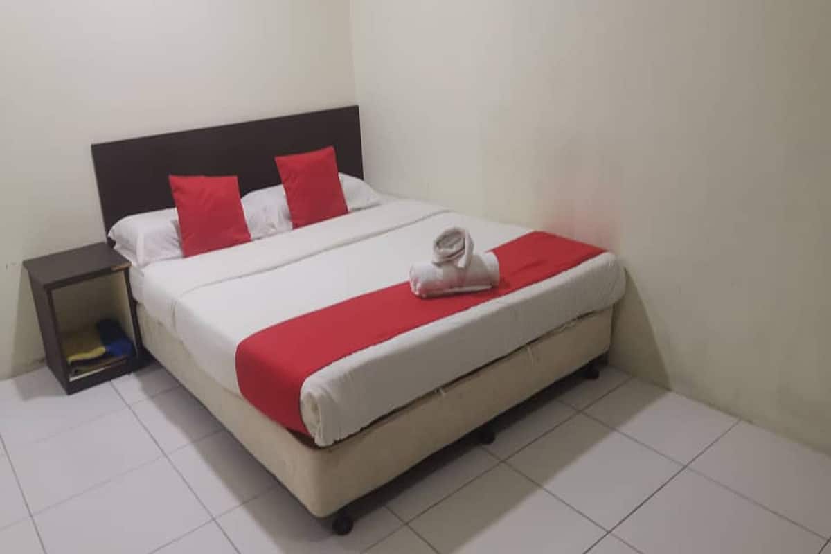 A neatly made bed with white and red bedding, a small bedside table with a lamp, and a plain white wall.