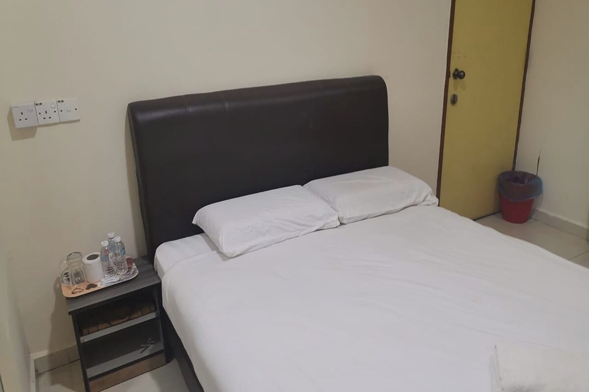 A single bed with white bedding, a dark headboard, a small bedside table with a tray of toiletries, and a yellow door in the background.