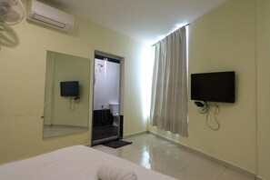 Habitación de hotel con cama, televisor montado en la pared, espejo, equipo de aire acondicionado y zona de ducha visible a través de una puerta abierta.