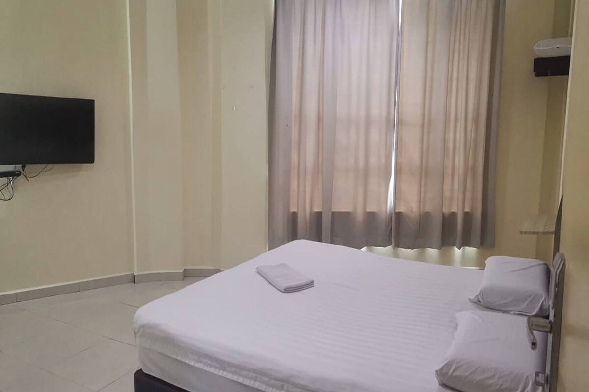 A neatly made bed with a white comforter and pillows, a flat-screen TV mounted on the wall, and a window with sheer curtains.