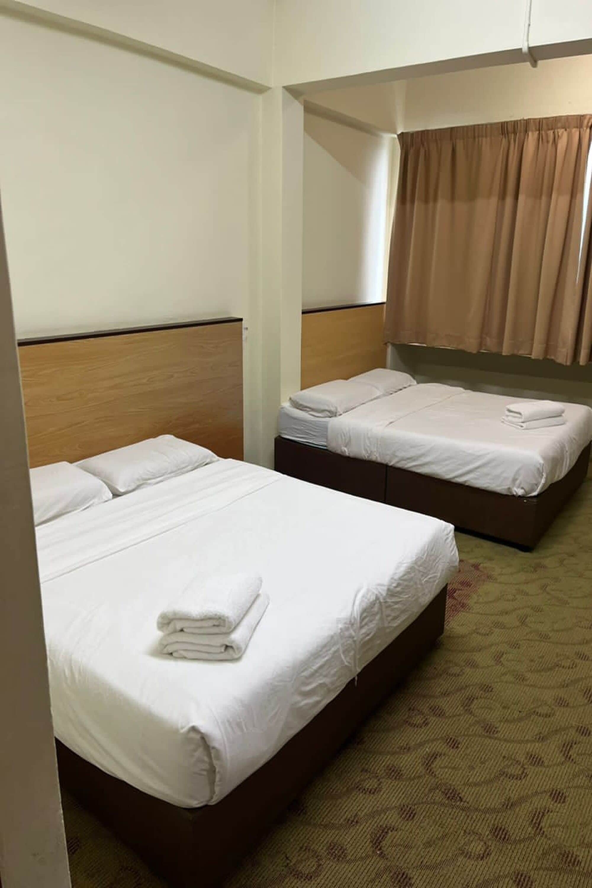 Habitación de hotel con dos camas individuales, cabecera de madera, cortina marrón y alfombra estampada.