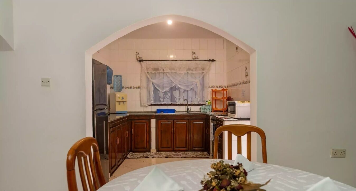 A dining area with a round table, chairs, and a kitchen visible through an archway.