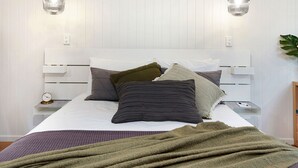 Un lit bien fait, avec une tête de lit blanche, plusieurs oreillers et une couverture verte. On y trouve deux tables de chevet, chacune avec une lampe, et une horloge sur l’une d’elles.