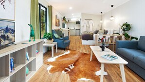 Un salon moderne avec un canapé, une table basse et un téléviseur. On aperçoit à l’arrière-plan un coin cuisine avec une table à manger et des chaises.