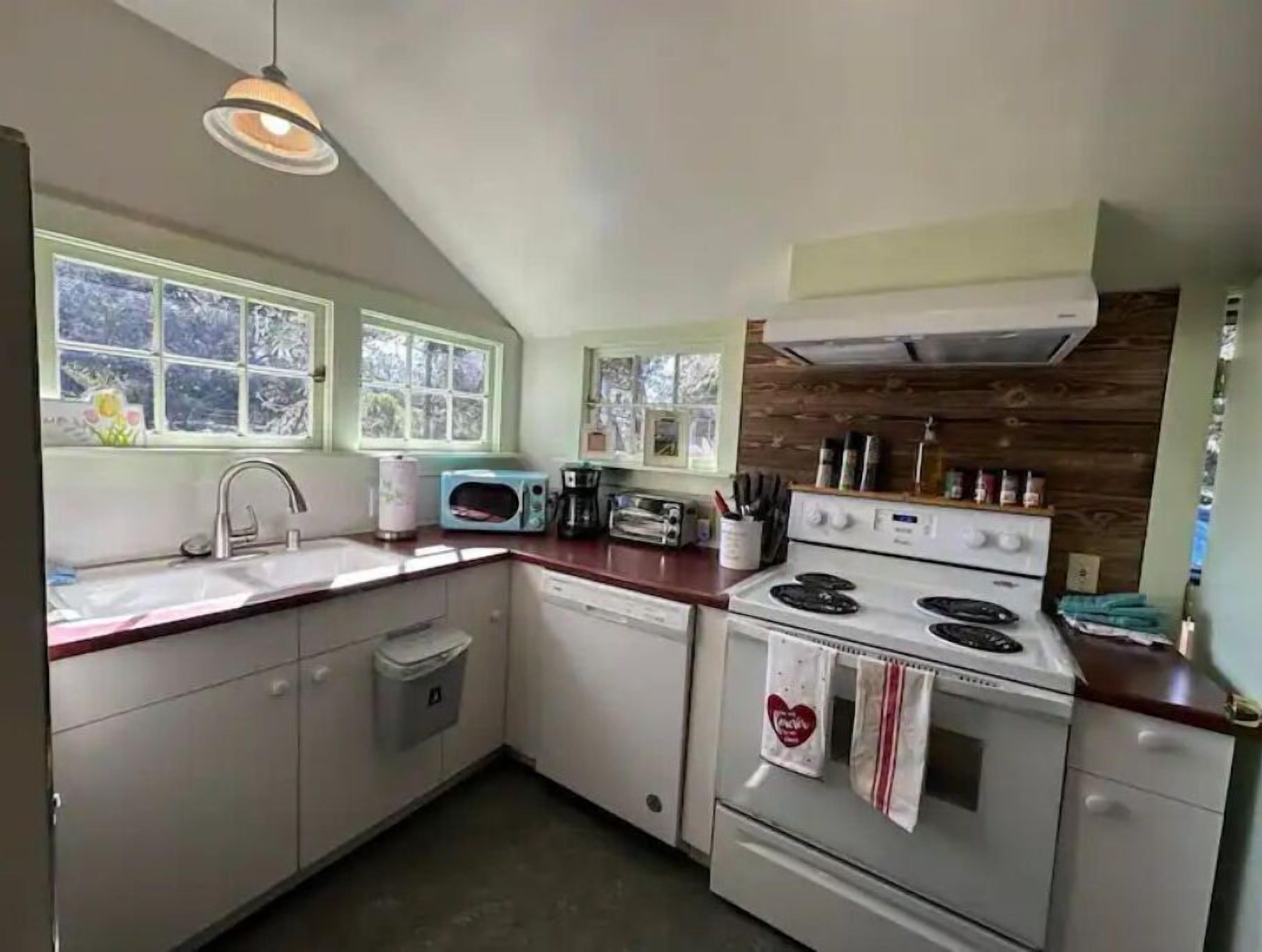 A compact kitchen with a sink, a microwave, a toaster, and a stove. There are windows providing natural light and a wooden accent wall.