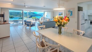 Une cuisine moderne avec des meubles blancs, un coin repas doté d’une table en verre et une vue sur la mer grâce à de grandes fenêtres.