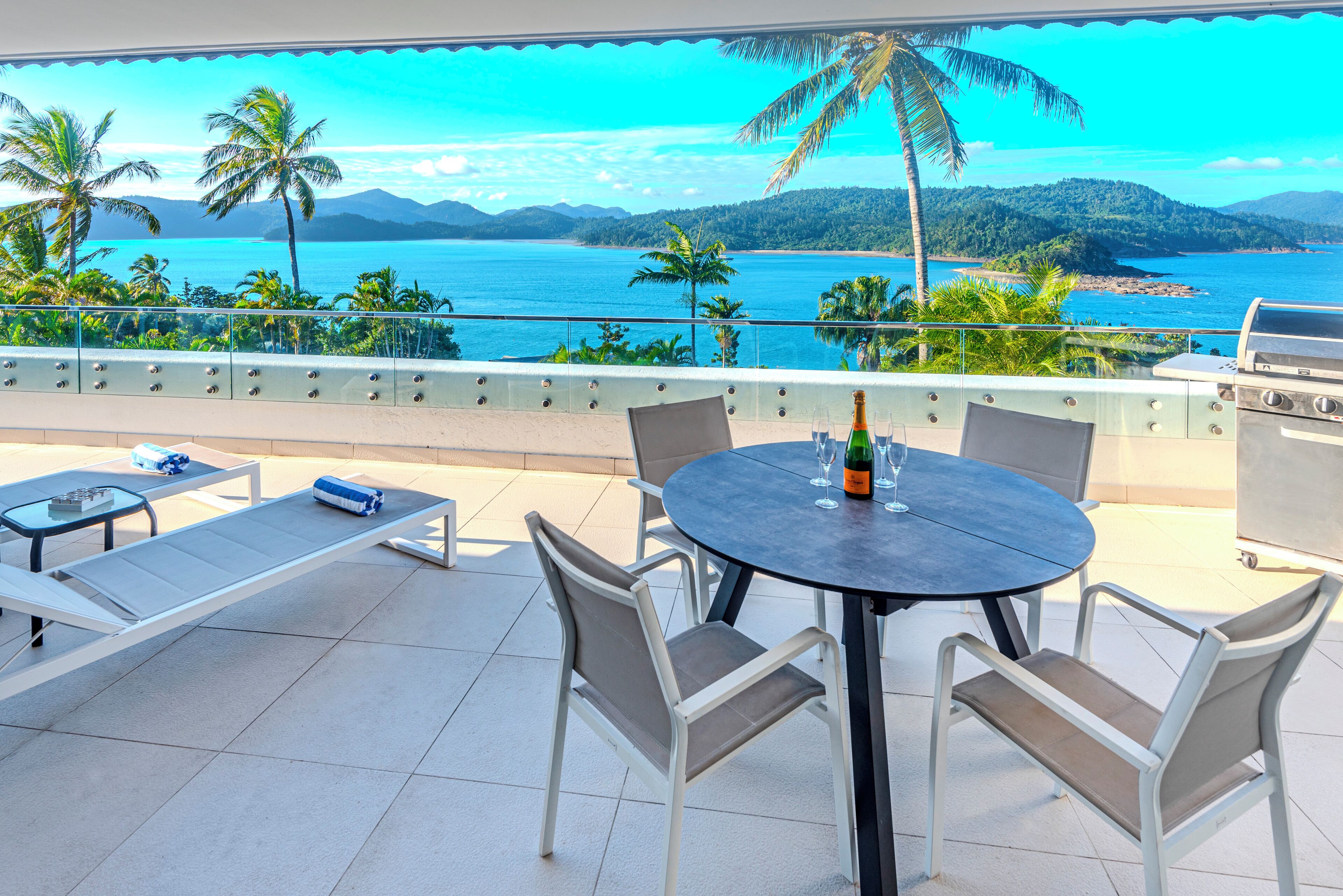 Un balcon avec vue sur l’océan, un barbecue et une table avec des chaises.